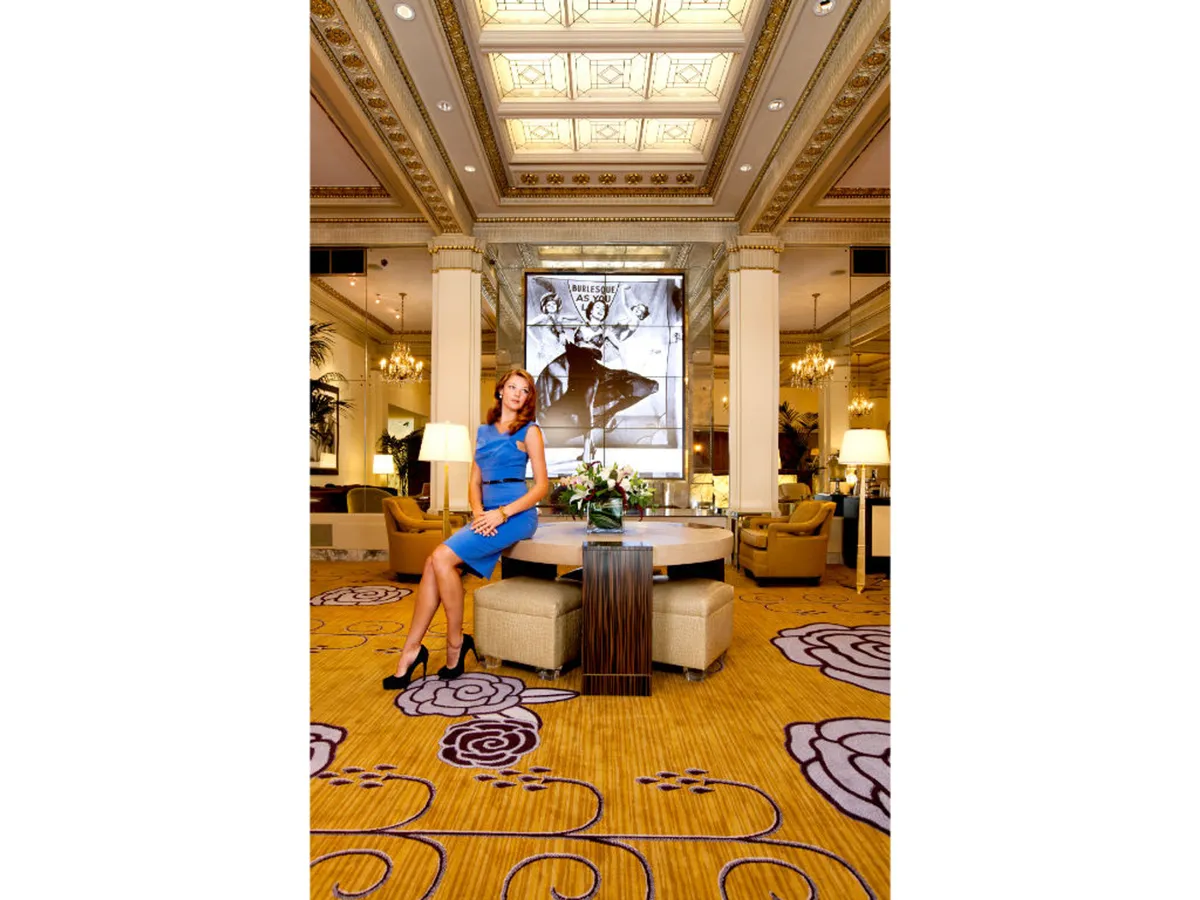 A sophisticated gathering space featuring a woman in a blue dress socializing near a lavish floral arrangement. The background showcases striking Golden Age film art, with other guests mingling in a luxurious, art-filled setting. The atmosphere is lively yet refined, emphasizing networking and stylish group interactions.
