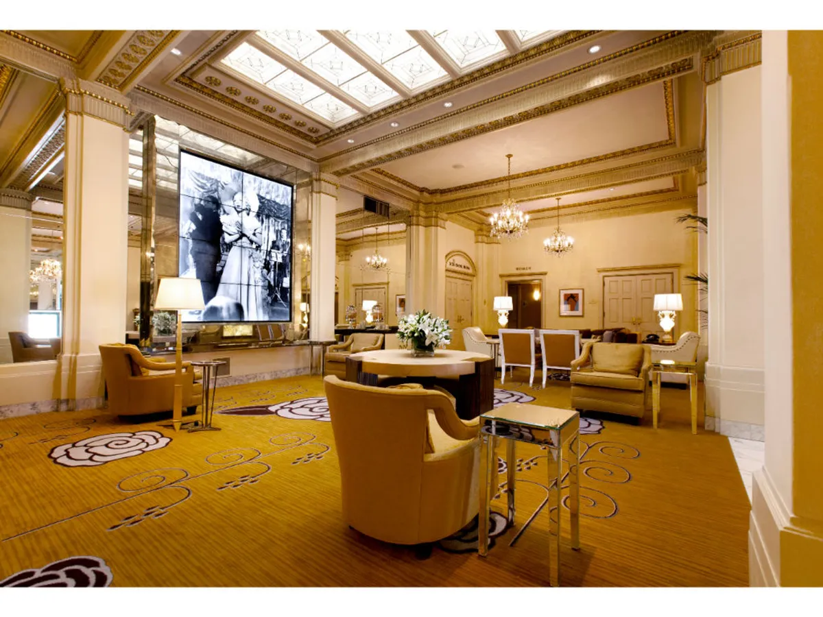 A spacious, elegant event room featuring a large digital screen ideal for presentations or screenings, illuminated by an ornate chandelier. The space has decorative carpeting, plush seating, and a central floral arrangement, emphasizing its suitability for gatherings, celebrations, or special events.