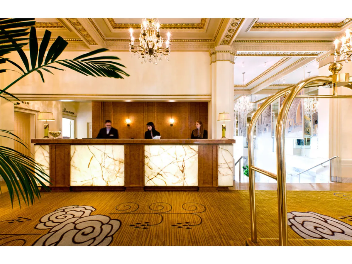 This image captures the reception area of the hotel, featuring a luxurious lobby with ornate chandeliers, a decorative carpet, and attentive staff members stationed at the front desk. The scene is lively and welcoming, with guests being greeted and engaging in conversation.
