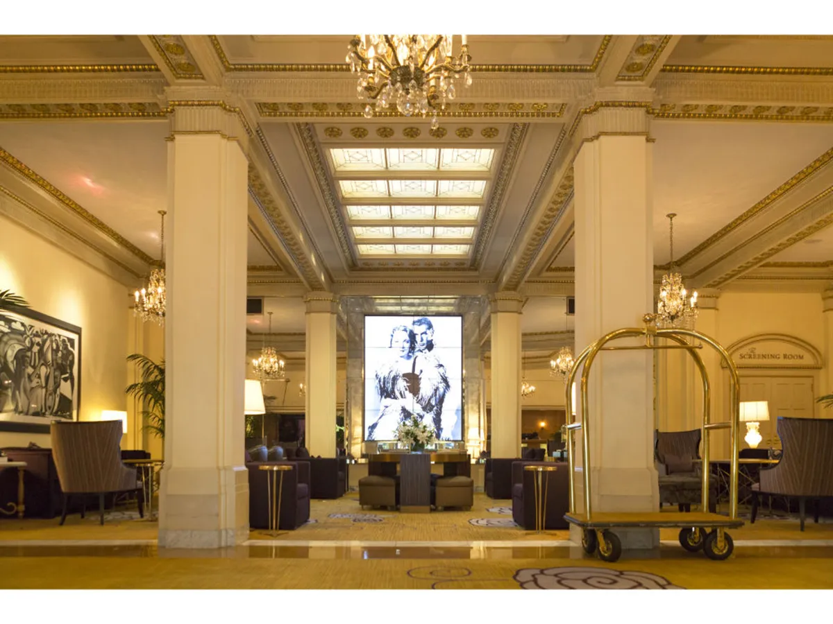 A spacious, sophisticated hotel lobby is shown, adorned with grand chandeliers, striking large-scale artwork, ample seating areas, and a visible luggage cart. The mood is both refined and inviting, with guests enjoying social and business moments in a well-appointed space.