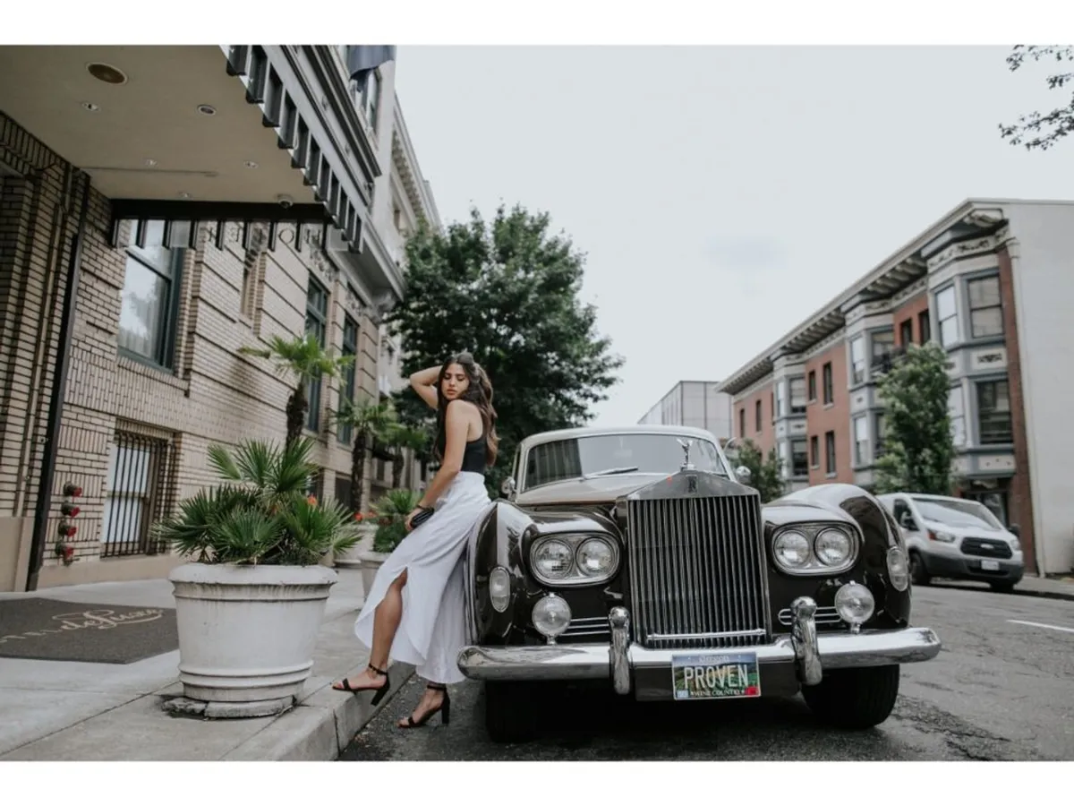 A vintage car is parked in front of an elegant building facade, with a woman in a white dress pausing gracefully nearby. Potted plants and urban architectural details frame the scene, evoking both glamour and a sense of exploration.