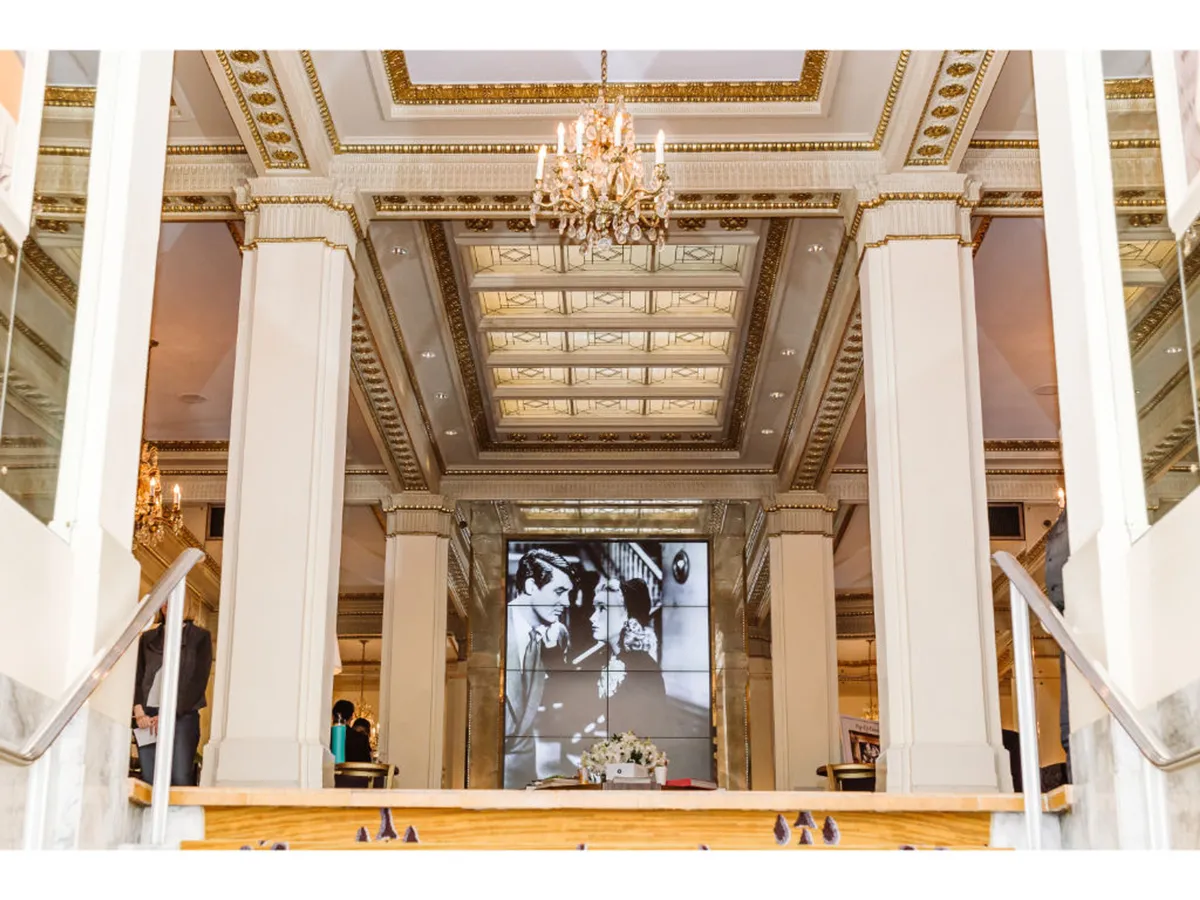 An elegant event room with a glittering chandelier, ornate decorative ceiling, and a large display screen. The setting is arranged for formal gatherings such as weddings or presentations, with an atmosphere of historic charm and sophistication. The room’s features suggest versatility for both intimate ceremonies and professional events.