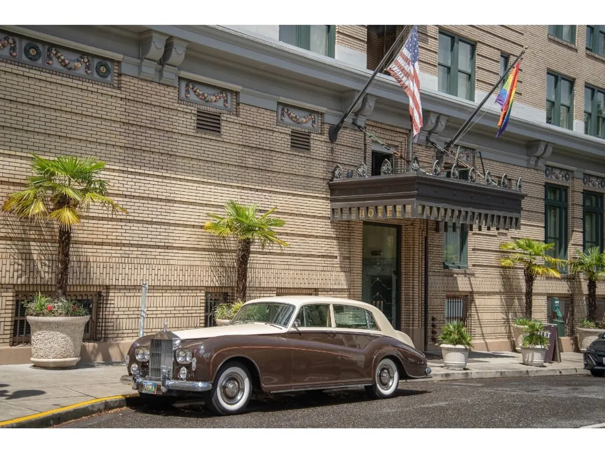 The exterior of the hotel is showcased with a classic Rolls-Royce parked at the ornate entrance, palm trees, and flags. The atmosphere is both luxurious and lively, evoking a sense of arrival and anticipation for city experiences.