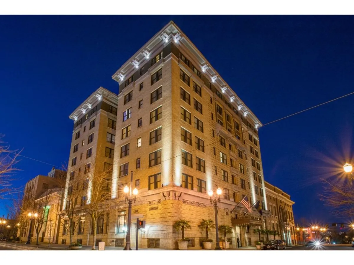 This image displays the ornate exterior of a sophisticated downtown building, illuminated by elegant light fixtures and framed by decorative palm trees and flag banners. The scene is lively, suggesting evening city exploration and the vibrant atmosphere of the surrounding neighborhood.