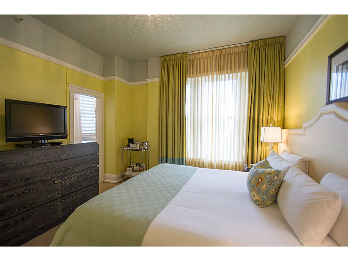 A sunlit Deluxe King room featuring a plush bed, elegant dresser, and inviting window seat framed by graceful curtains. Morning light pours through the window, illuminating the cozy space ideal for relaxing, reading, or enjoying a peaceful coffee while watching city life unfold below.
