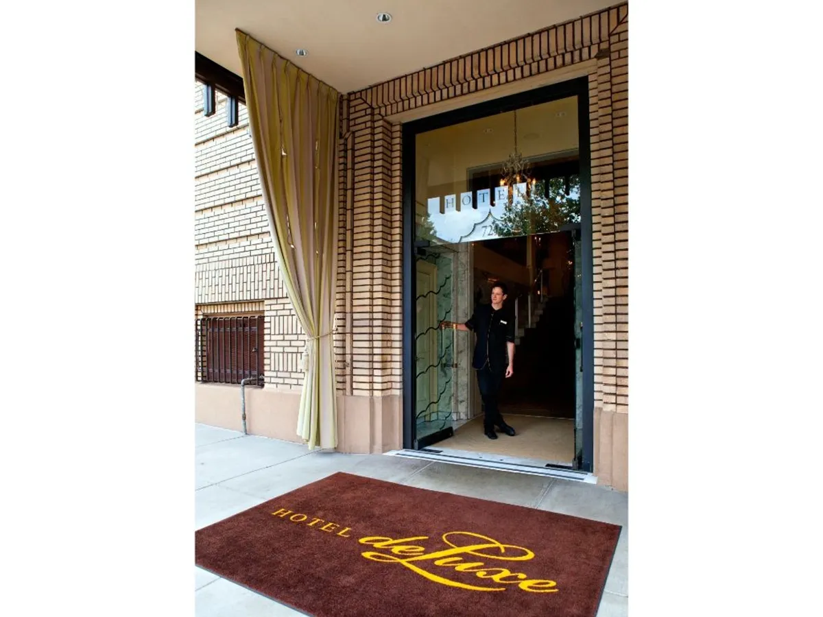 This image captures the elegant exterior entrance of the hotel, featuring a welcoming staff member, the hotel name mat, and a sophisticated façade. The scene suggests a personalized and inviting arrival experience for guests.