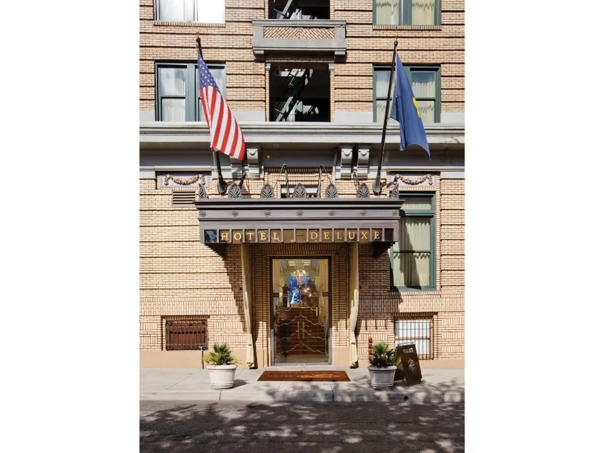The grand entrance of Hotel Deluxe is showcased with large glass doors, the hotel’s name prominently displayed above, and flags waving overhead. The scene conveys a sense of arrival and elegant anticipation.
